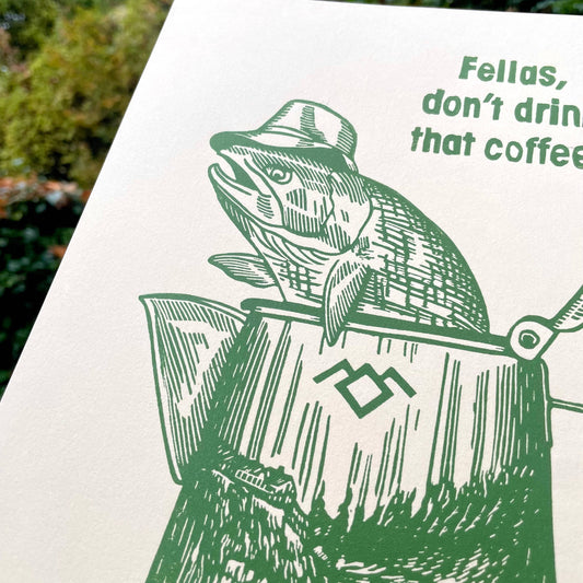 Print with a fish wearing a hat in a coffee percolator and text 'Fellas, don't drink that coffee!' held against a natural background.