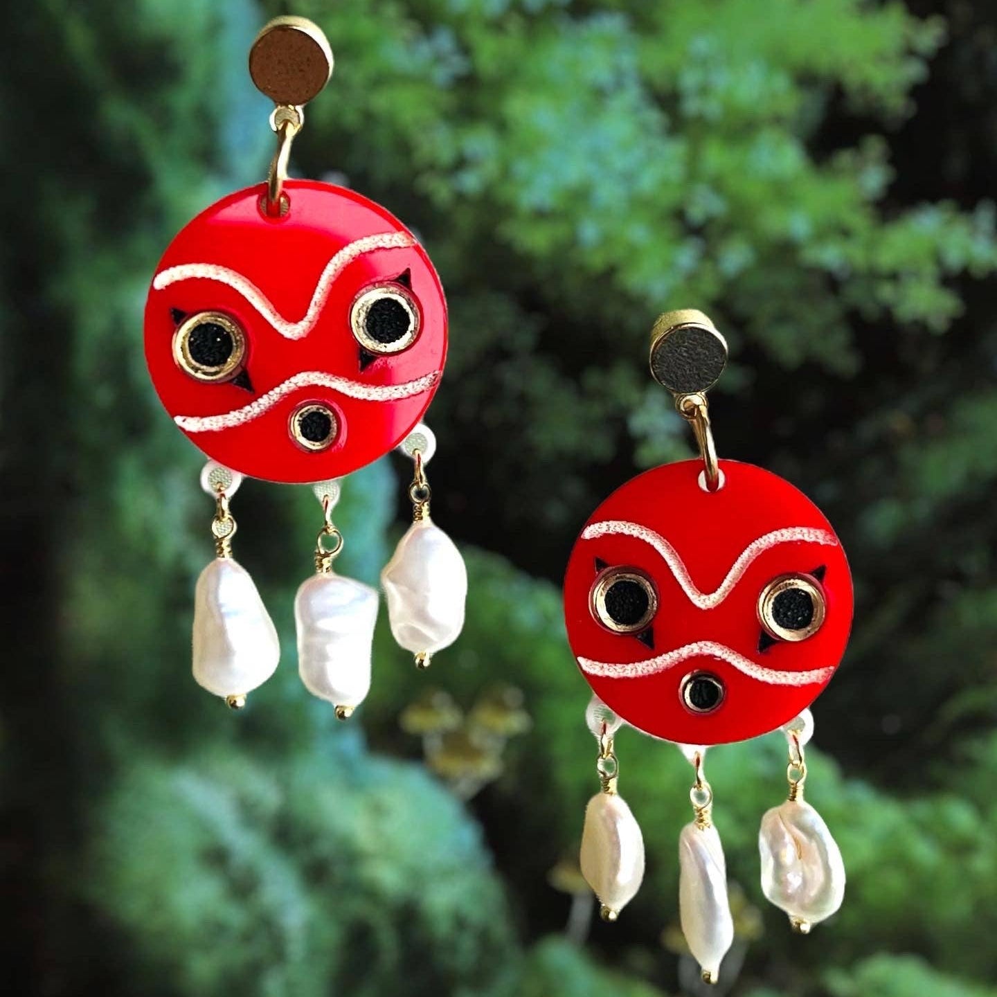 Red mask earrings based on San's wolf mask from the Studio Ghibli movie, Princess Mononoke, with white freshwater pearl beads against a green foliage background