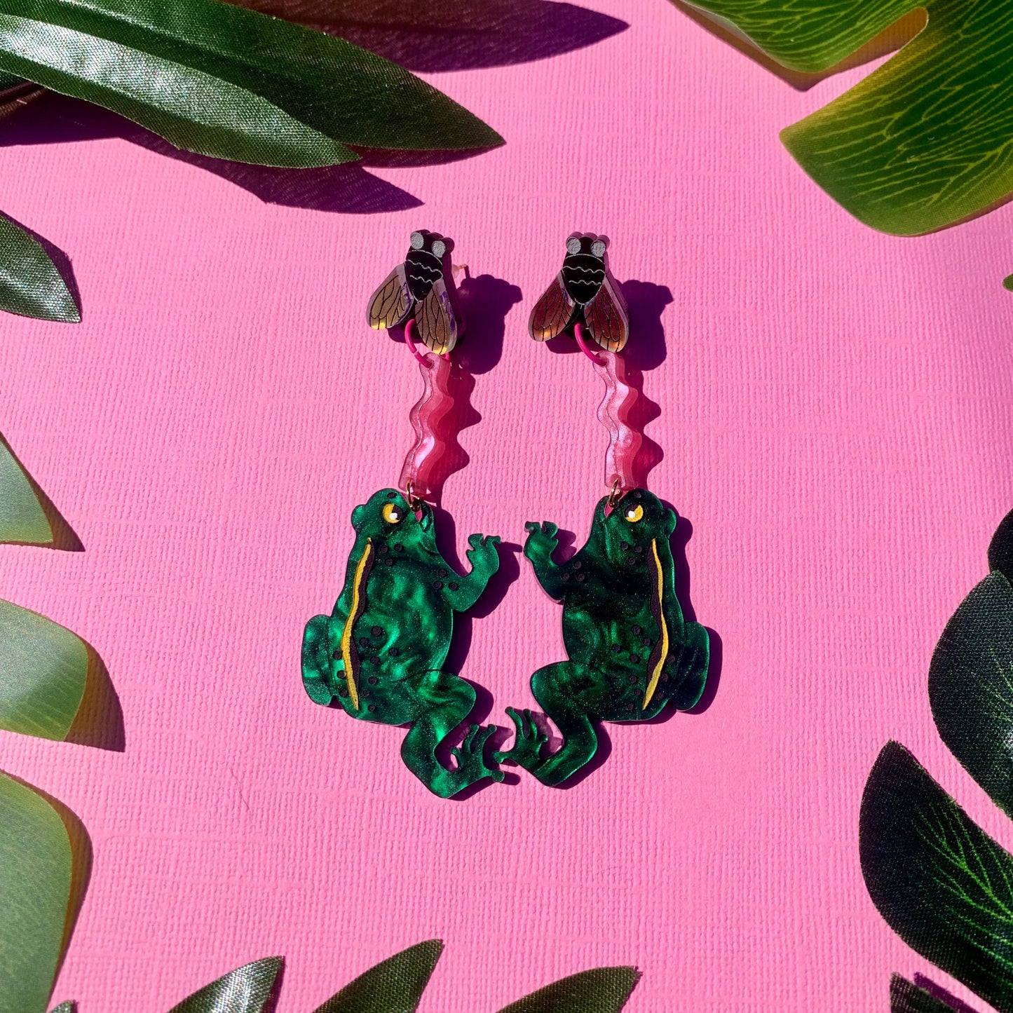 Frog-shaped earrings with flies on a pink background with green leaves