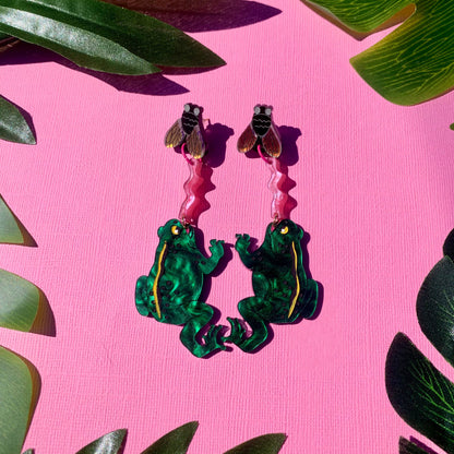 Frog-shaped earrings with flies on a pink background with green leaves