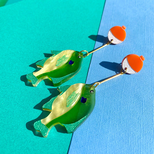 Green fish-shaped earrings with orange floats on a blue and green background