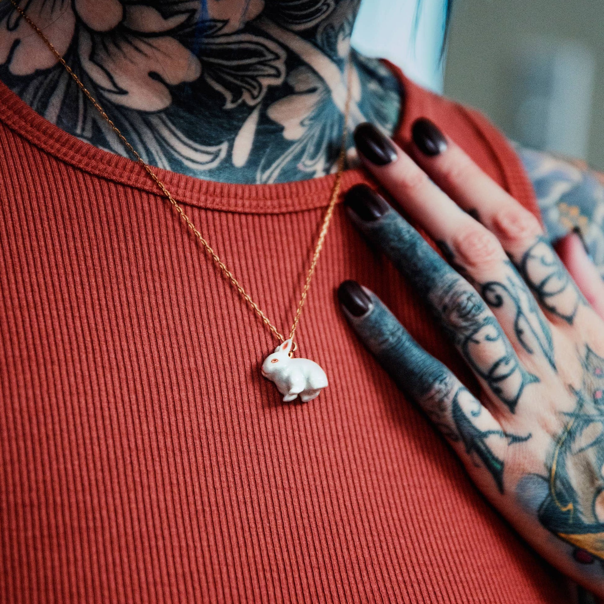 Person with tattoos wearing a red ribbed shirt and gold necklace with a bunny pendant.