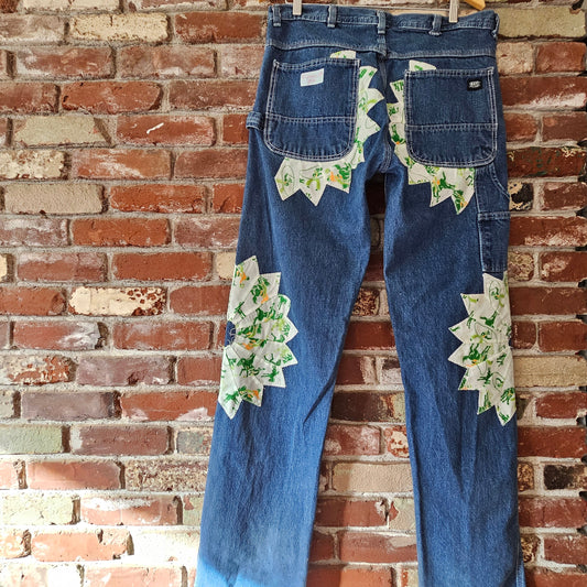 Blue jeans with rodeo patches on a wooden hanger against a brick wall.