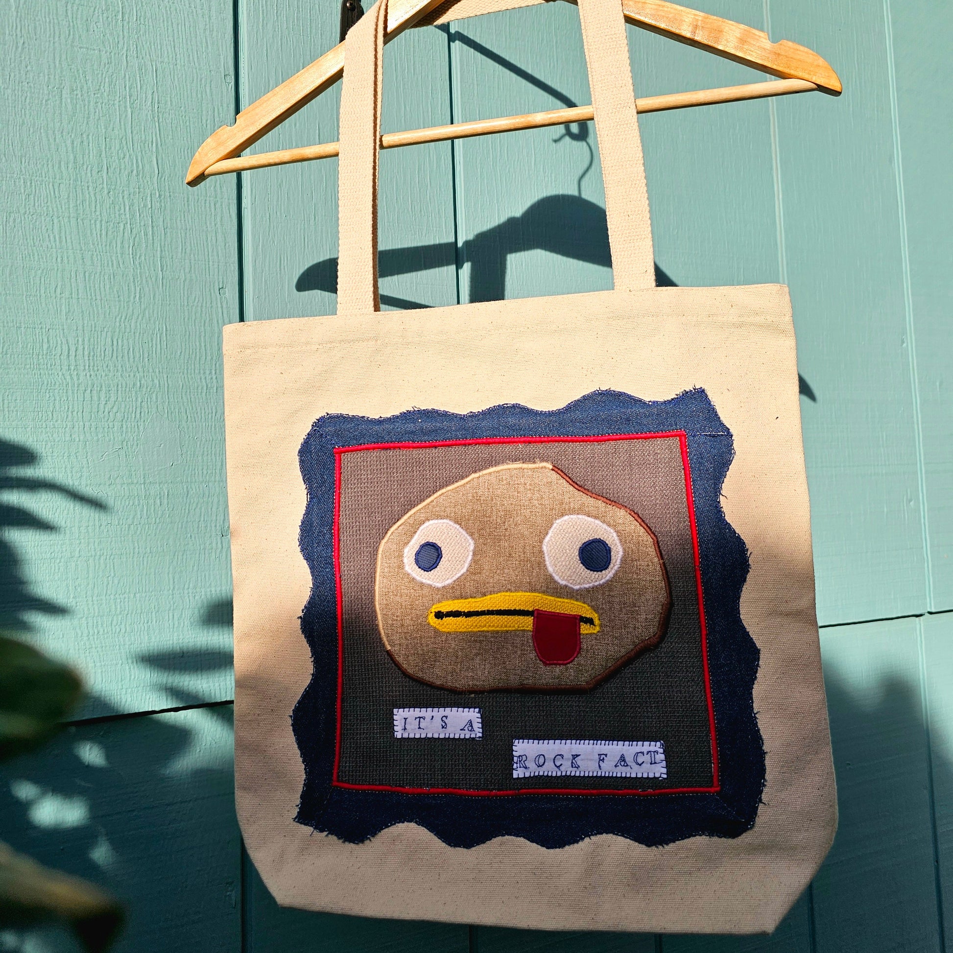 Tote bag with rock fact design from Over the Garden Wall on a wooden hanger against a teal wall.