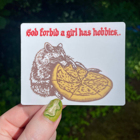 Stricker with a raccoon eating a pizza with text above reading "God forbid a girl has hobbies"  held by a hand against a blurred green background