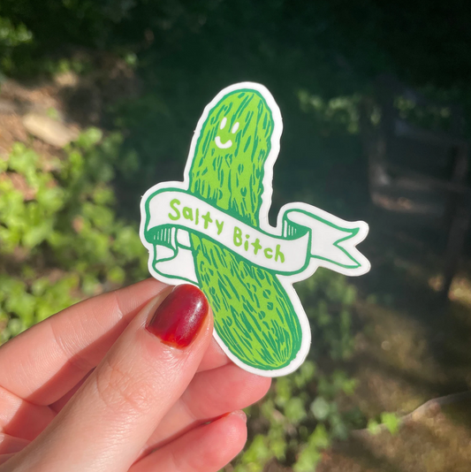 Sticker of a pickle with 'Salty Bitch' text held by a hand against a blurred natural background