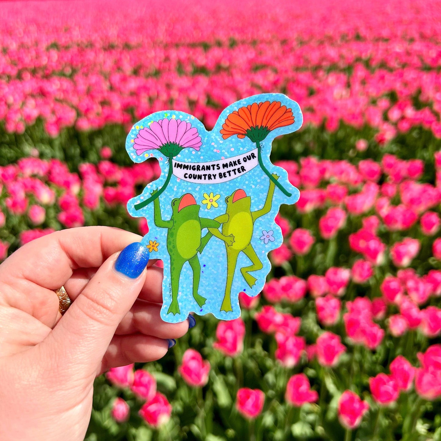 Hand holding a colorful sticker with two frogs and text that says "immigrants make our country better" in front of pink flowers