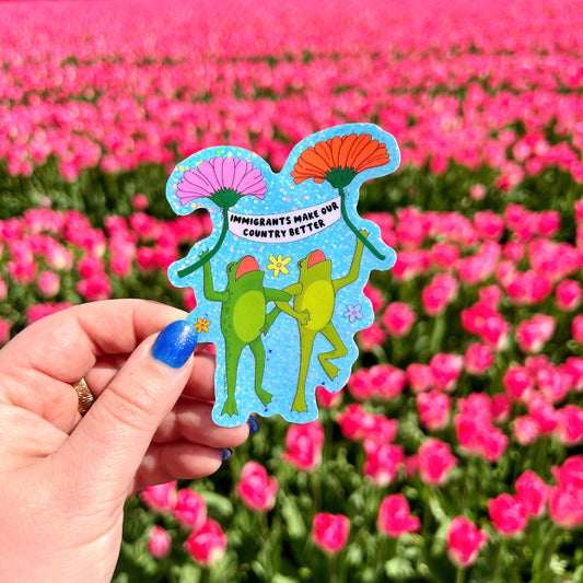 Hand holding a colorful sticker with two frogs and text that says "immigrants make our country better" in front of pink flowers