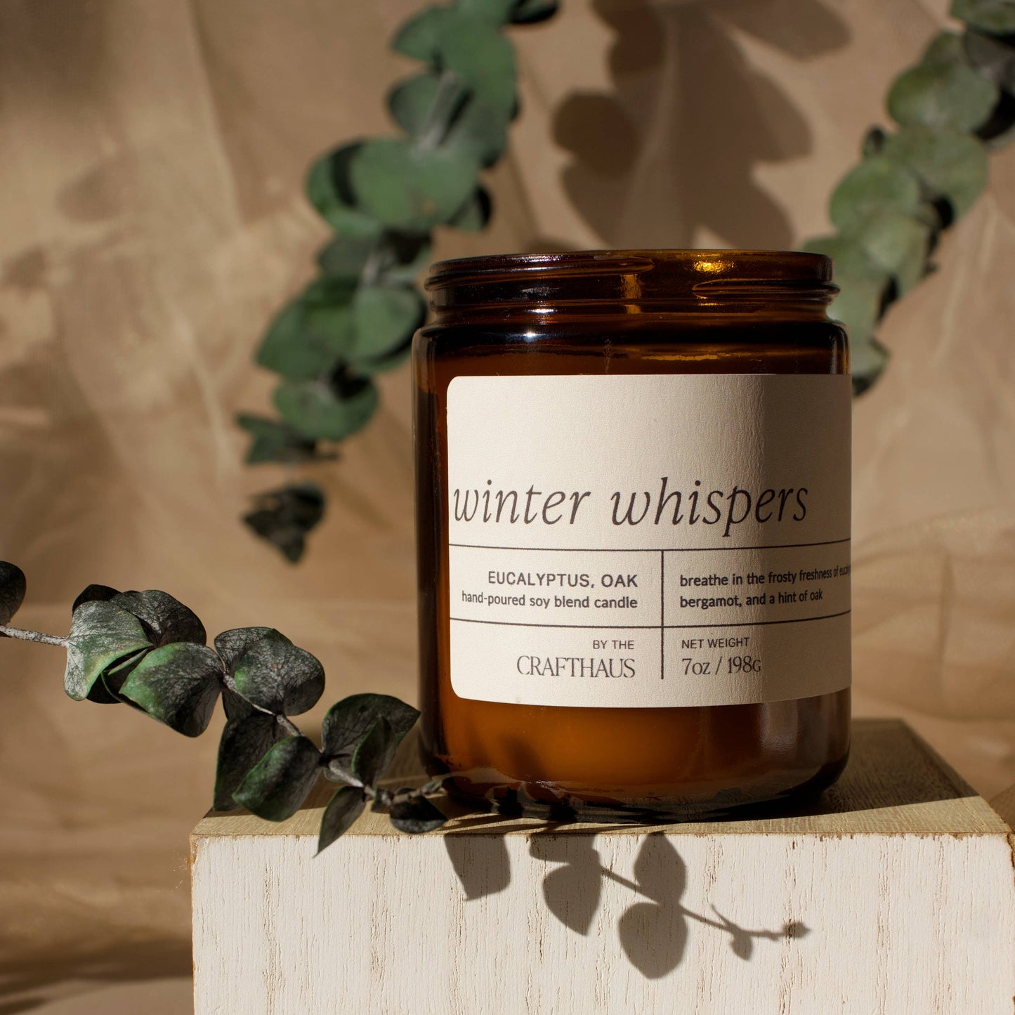 Candle labeled 'winter whispers' on a wooden block with eucalyptus leaves.