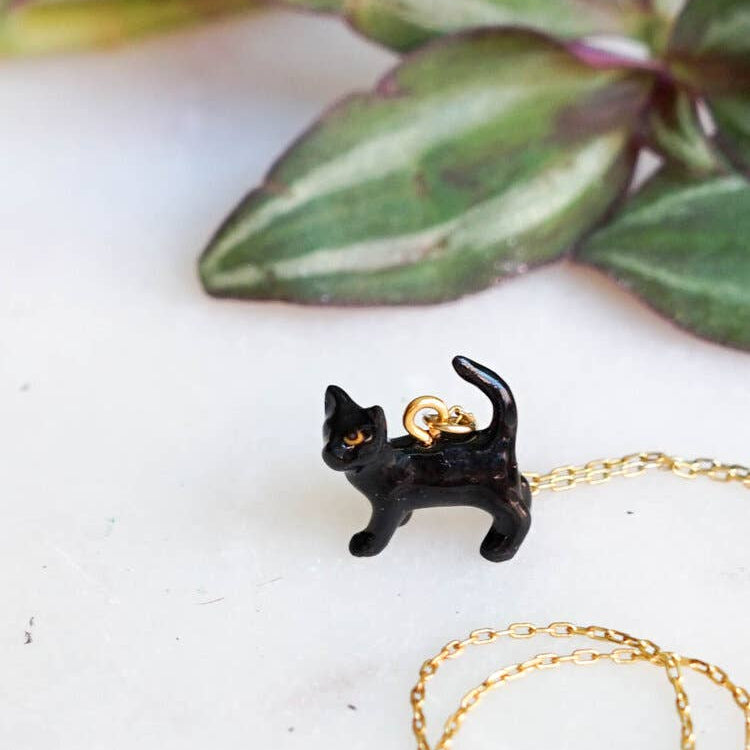 Black cat-shaped pendant on a gold chain with green leaves in the background