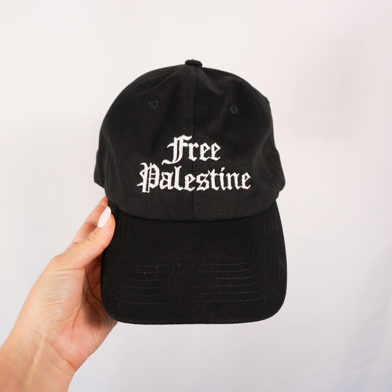 Black cap with 'Free Palestine' text held by a hand against a light gray background