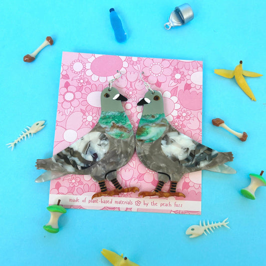 Pair of pigeon bird earrings on a pink card with trash accessories surrounding them on a blue background