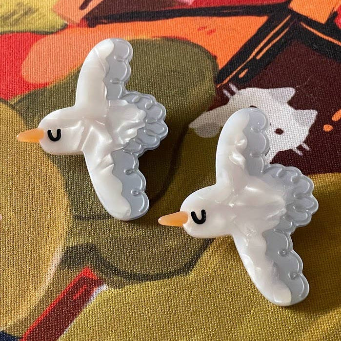Two white bird-shaped earrings on a colorful fabric background