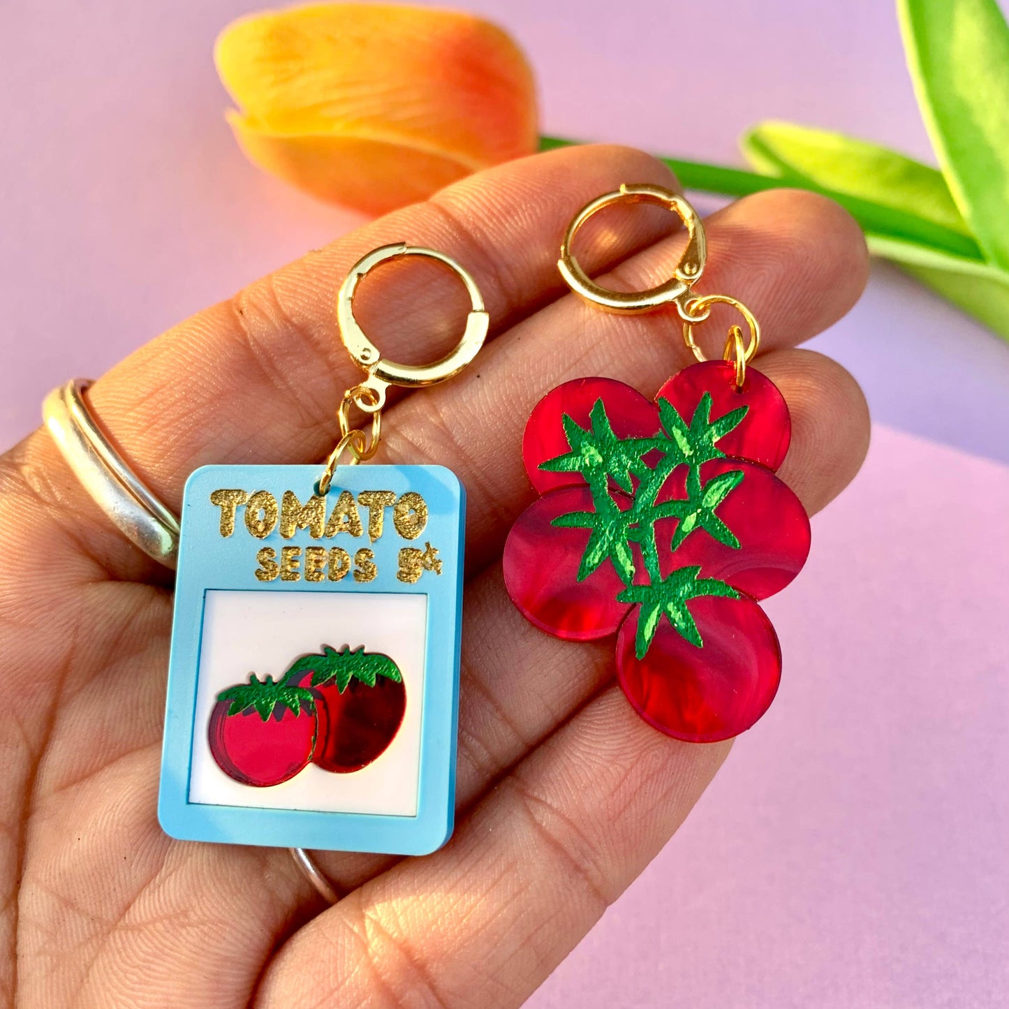 Hand holding two earrings: one resembling a seed packet with tomatoes and the other a red tomato flower, against a light purple background.