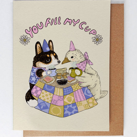 Greeting card with a dog and duck at a table, both holding a mug, with 'You Fill My Cup' text above.