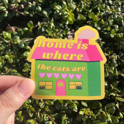 House-shaped sticker with 'home is where the cats are' text held in front of green foliage