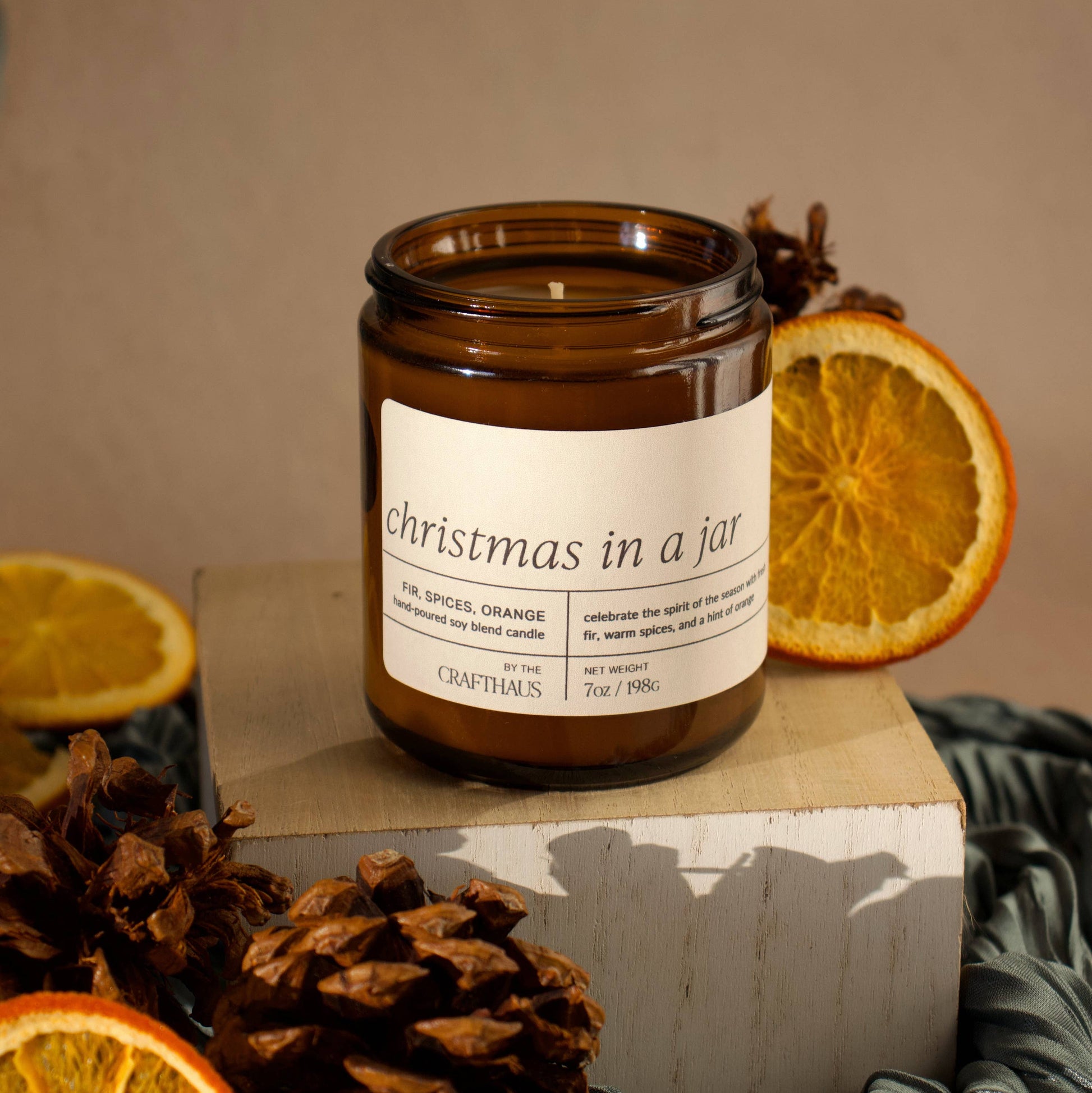 Candle labeled 'Christmas in a Jar' surrounded by oranges and pine cones on a neutral background