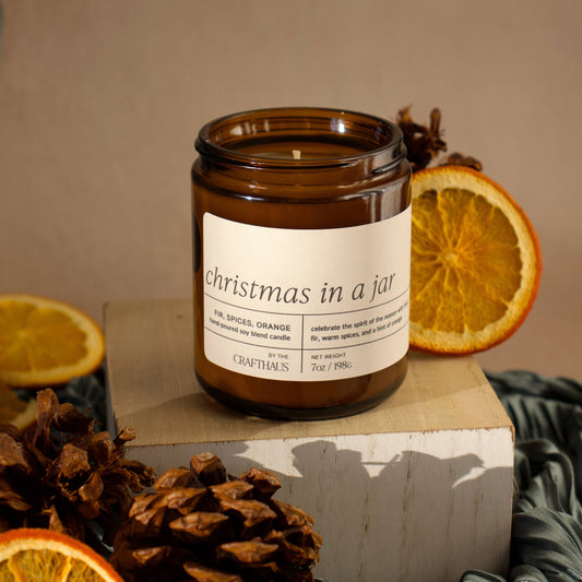 Candle labeled 'Christmas in a Jar' surrounded by oranges and pine cones on a neutral background