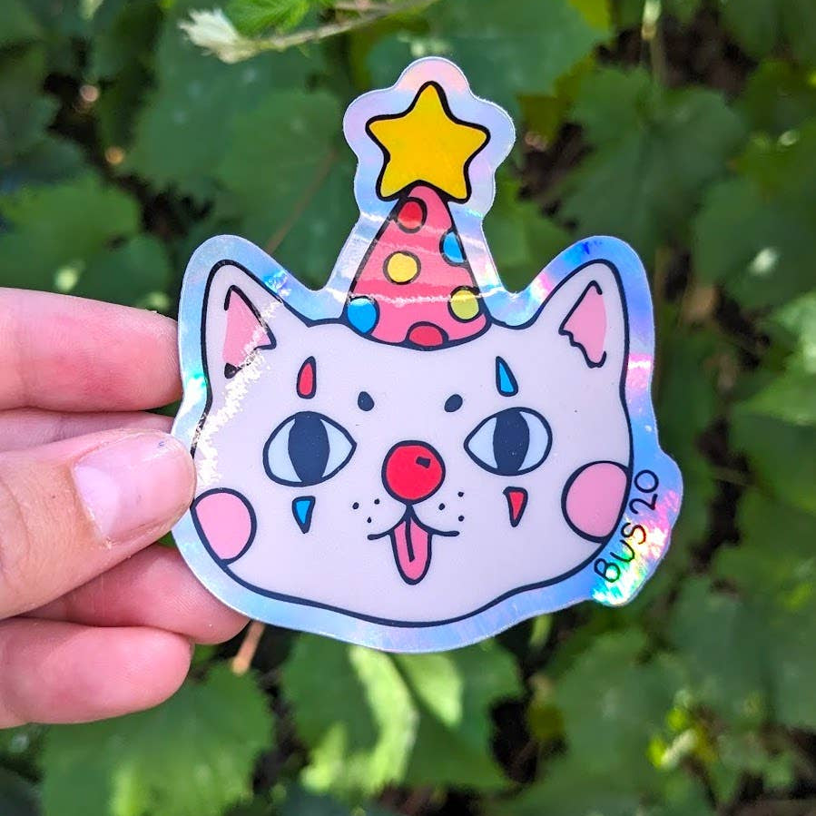 Holographic sticker of a cat with a party hat held against a green leafy background