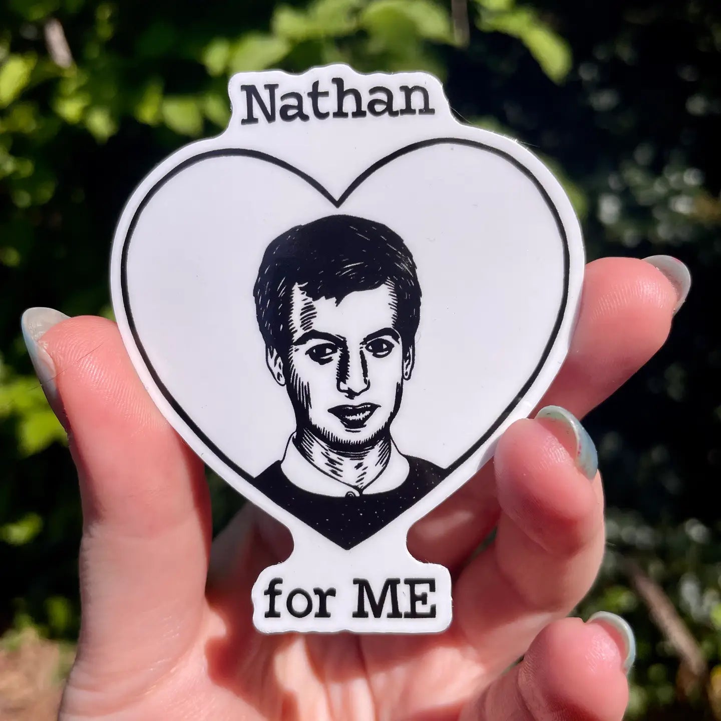 Heart-shaped sticker with a portrait and 'Nathan for ME' text held in a hand against a blurred green background