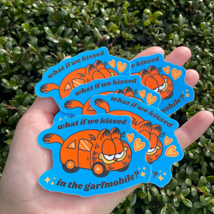 Sticker with the Garfield Mobile that reads "What if we kissed in the garfmobile?" in someone's hand in front of green foliage