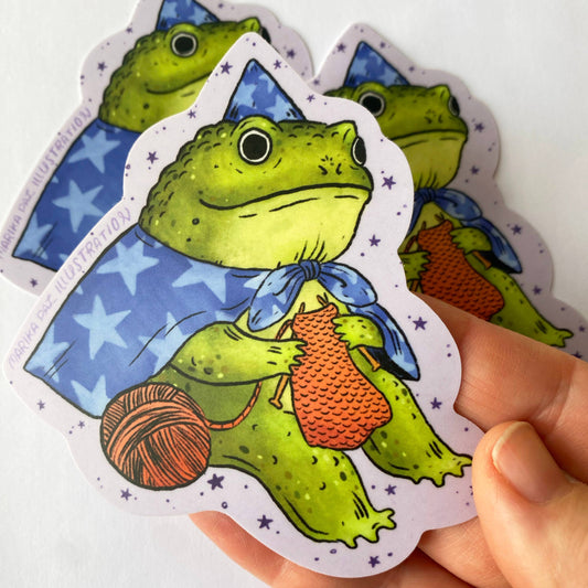 Hand holding a sticker of a green frog with yarn and knitting needles against a white background