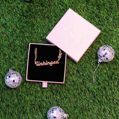 Gold necklace with 'Unhinged' on a black box, surrounded by disco balls on grass