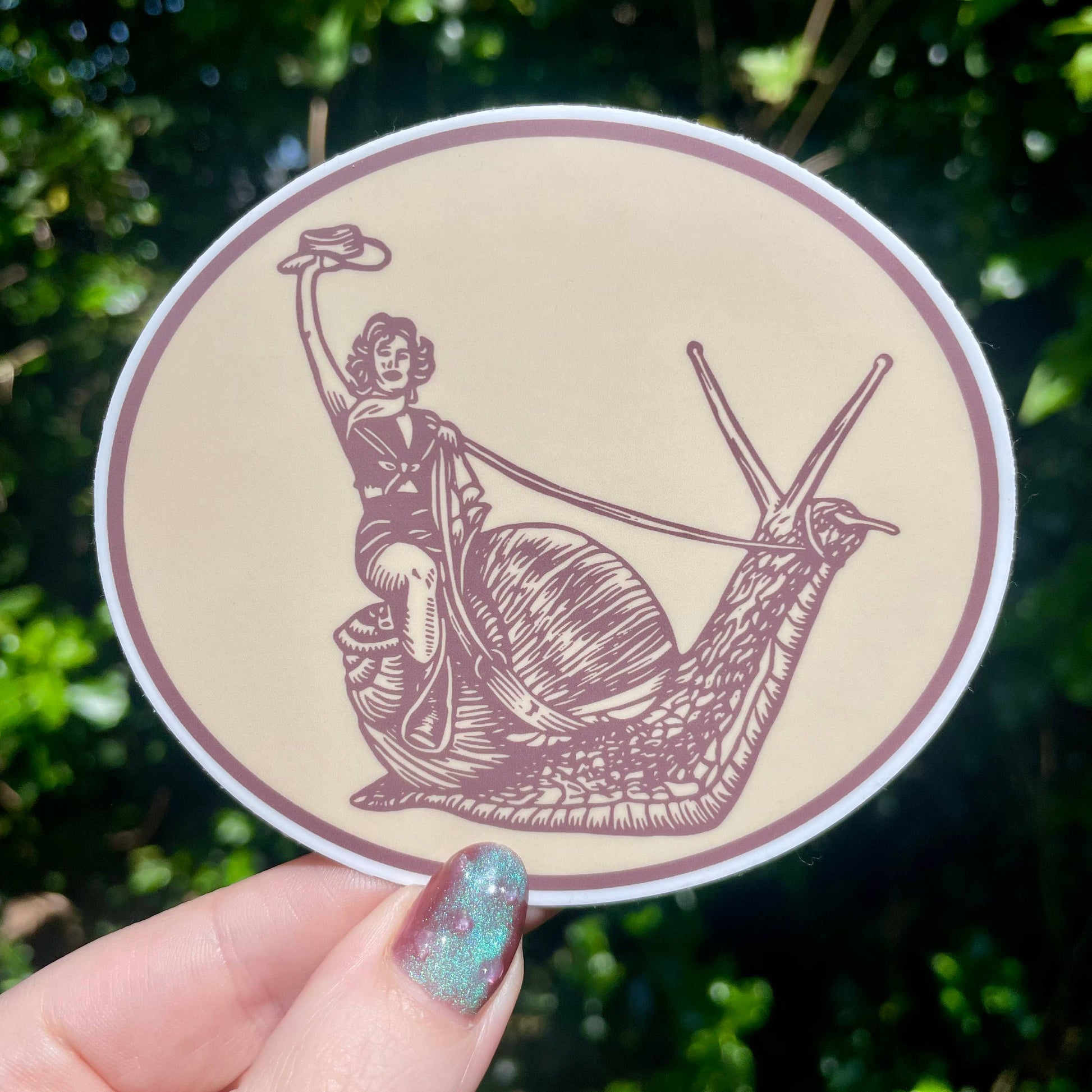 Circular sticker with a design of a person riding a snail, held by a hand against a green leafy background.