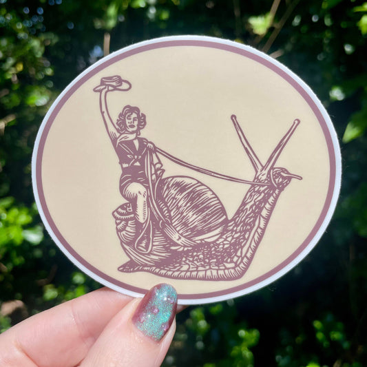 Circular sticker with a design of a person riding a snail, held by a hand against a green leafy background.