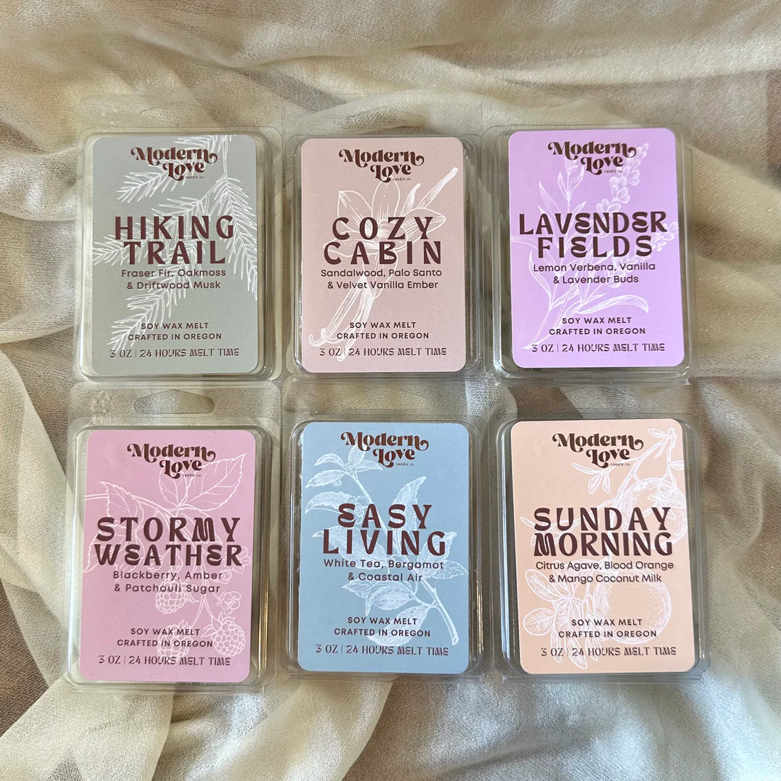 Set of six small rectangular soy wax melt packages with different scents on a beige fabric background