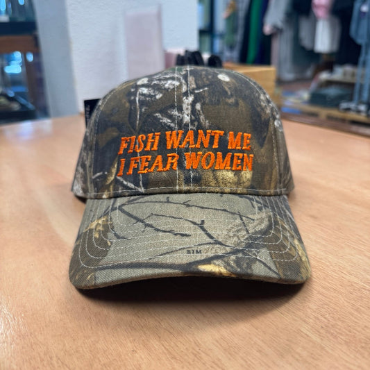 A camouflage baseball cap with orange embroidered text reading 'FISH WANT ME I FEAR WOMEN' sits on a wooden table inside a retail store.