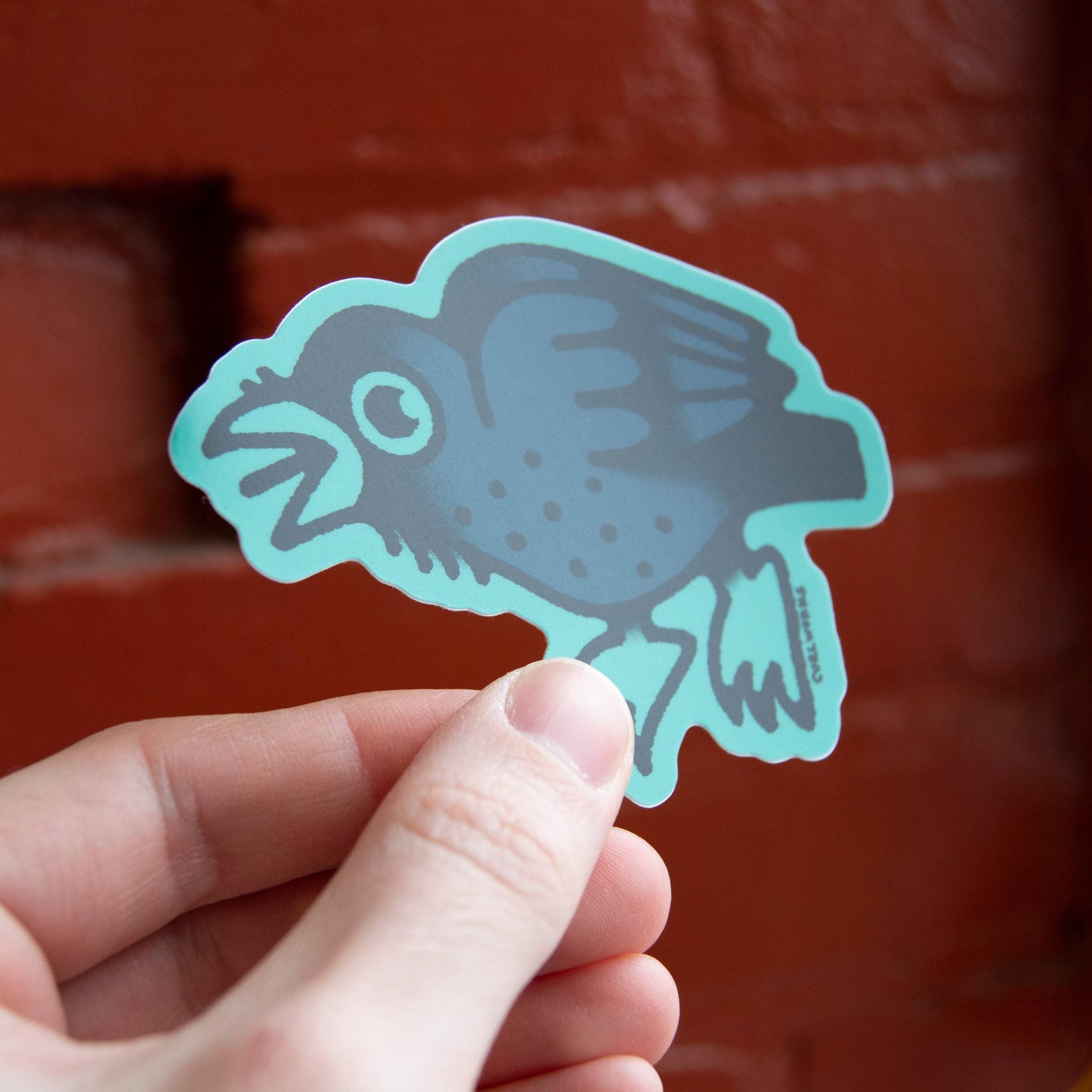 Hand holding a teal sticker of a crow against a red brick wall.