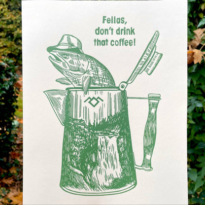 Print with a fish wearing a hat in a coffee percolator and text 'Fellas, don't drink that coffee!' held against a natural background.
