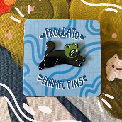 Frog-hat wearing black cat enamel pin on a blue card with 'Froggato' branding against a colorful background.