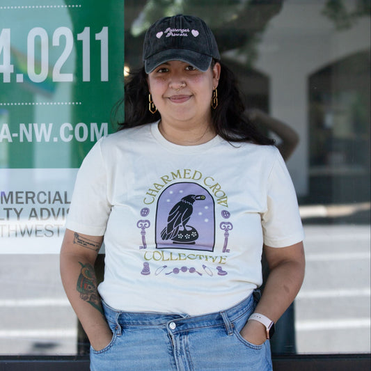 Person wearing a beige Charmed Crow t-shirt with a graphic design and denim shorts standing outdoors. The arch and trinkets in the design are lavender, the text is green.
