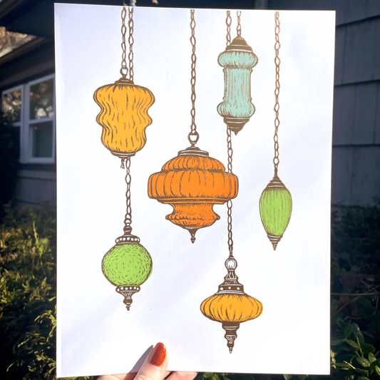 Colorful hanging lamps printed on a white piece of paper held by a hand outdoors.