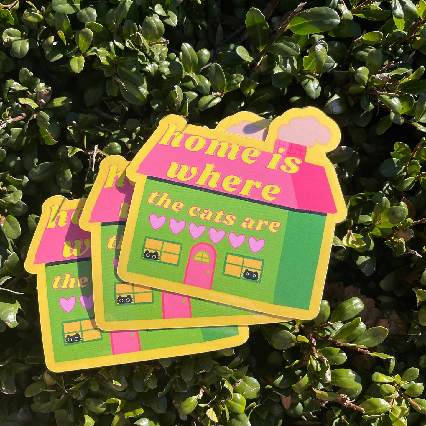 Three colorful stickers with a house design and text 'home is where the cats are' on a green leafy background.