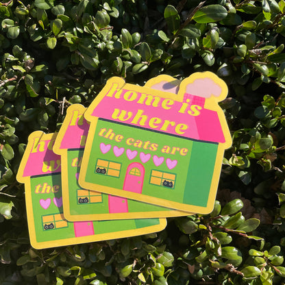 Three colorful stickers with a house design and text 'home is where the cats are' on a green leafy background.