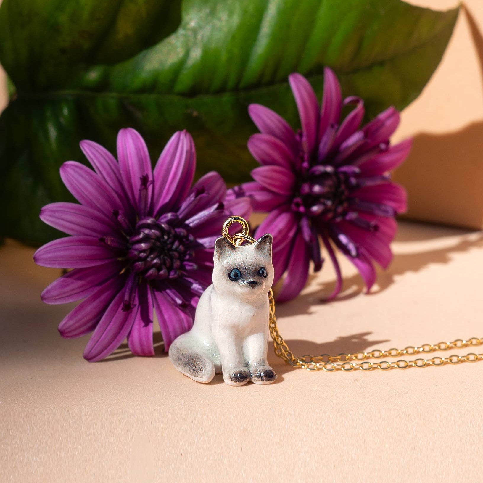 Cat-shaped pendant on a chain with purple flowers and green leaves in the background