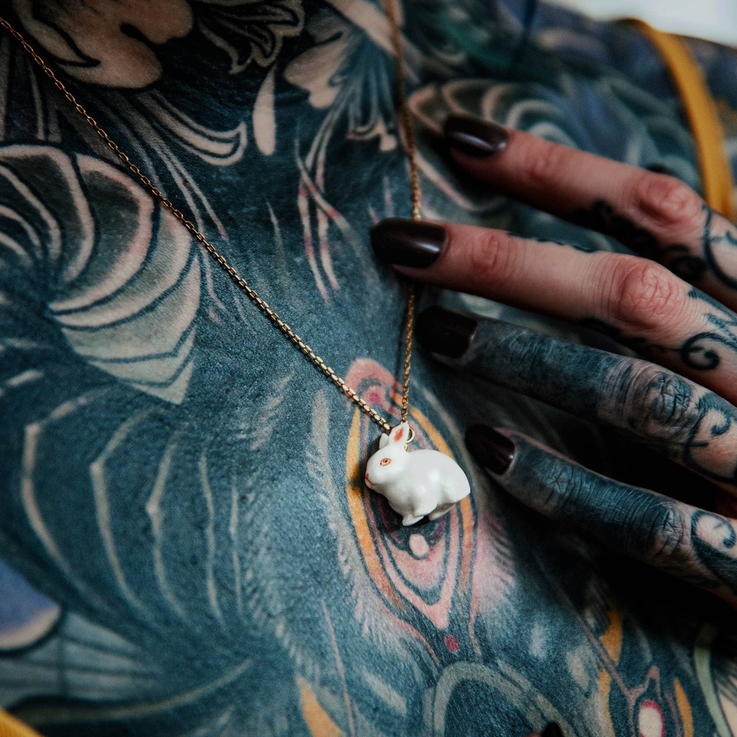 Close-up of a tattooed person with hand their hands touching a necklace with a small white bunny pendant on their neck