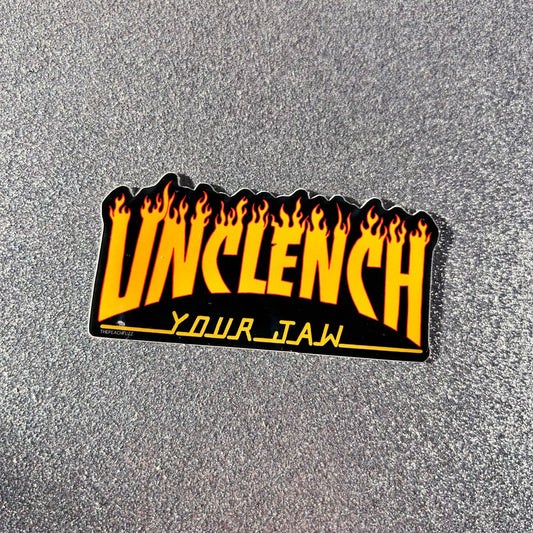 A rectangular enamel pin with the word 'UNCLENCH' in bold yellow letters outlined in black, featuring orange and yellow flames along the top edge, and 'YOUR_JAW' in yellow at the bottom, displayed on a textured gray background.