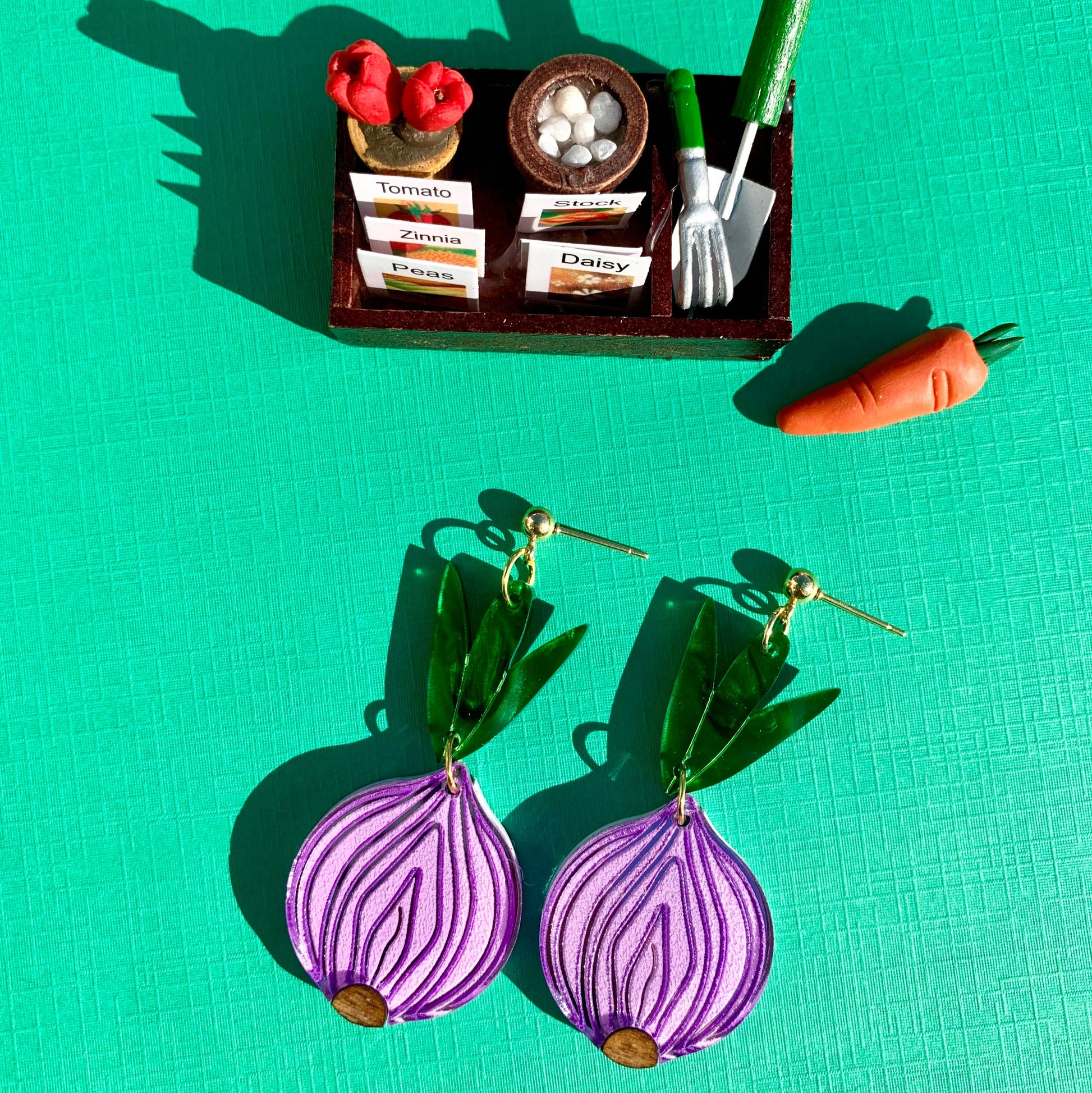 Purple onion-shaped earrings on a green background with miniature gardening set.