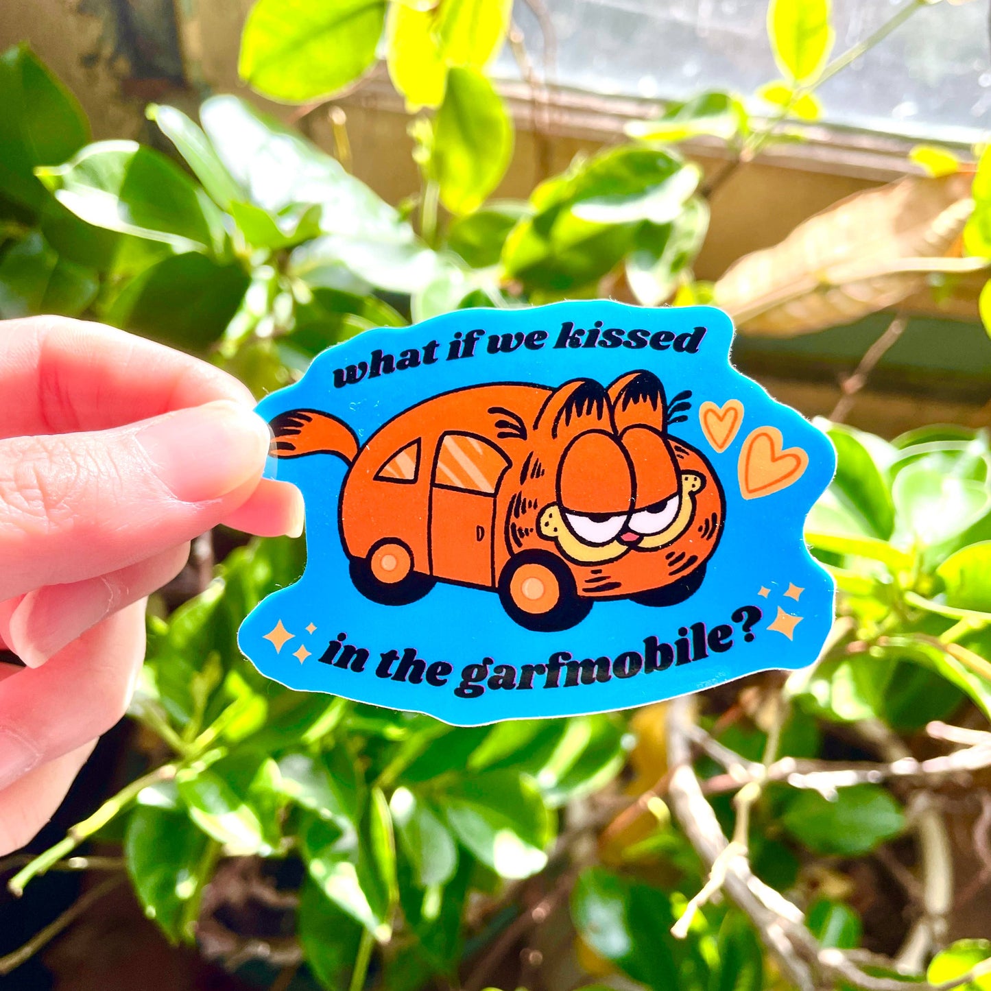 Sticker with the Garfield Mobile that reads "What if we kissed in the garfmobile?" held in front of a plant